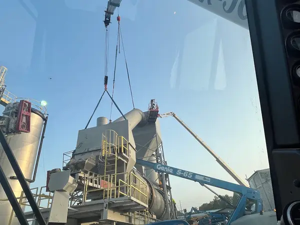 The view from inside a Gordon Crane cab at first light, looking out through the glass past the load line to a plant structure hanging in slings, with the operator's phone number lettered along the boom.