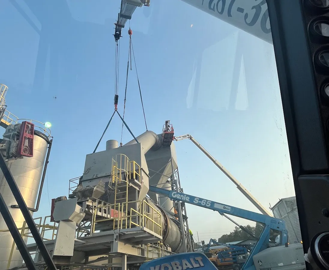 The view from inside a Gordon Crane cab at first light, looking out through the glass past the load line to a plant structure hanging in slings, with the operator's phone number lettered along the boom.