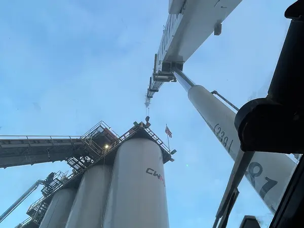 Looking straight up through a Gordon Crane windscreen at dusk, the white boom running out overhead toward a row of tall plant silos with a flag flying from the platform.