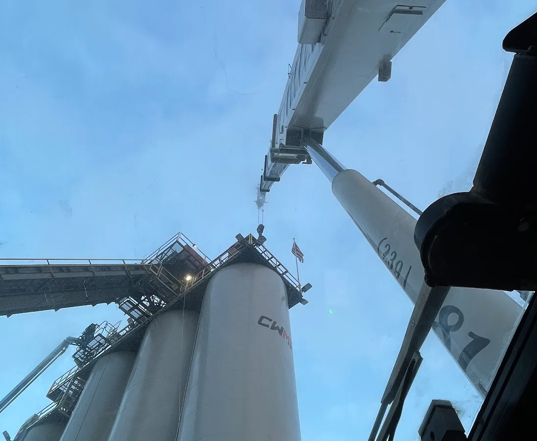 Looking straight up through a Gordon Crane windscreen at dusk, the white boom running out overhead toward a row of tall plant silos with a flag flying from the platform.