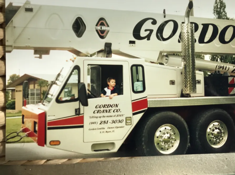 A young boy sitting in the cab of a Gordon Crane truck crane, the door lettered “Gordon Crane Co. — Lifting up the name of JESUS — (941) 281-3030 — Gordon Centilla, Owner/Operator, N. Ft. Myers, Fl”.
