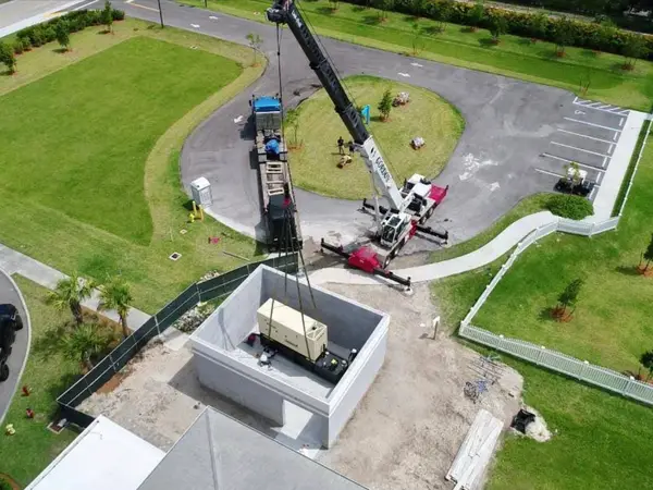 Aerial view of a Gordon Crane machine set up on outriggers, lowering a generator into a walled concrete enclosure beside a newly finished parking lot.