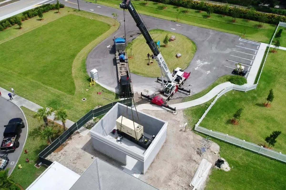 Aerial view of a Gordon Crane machine set up on outriggers, lowering a generator into a walled concrete enclosure beside a newly finished parking lot.