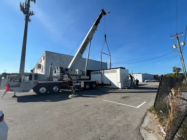 Gordon Crane's boom rigged over a long white packaged air-conditioning unit on a lowboy trailer, its row of condenser fans along the top and louvred coil panels down the side, with two crew on ladders working alongside it at a commercial building.