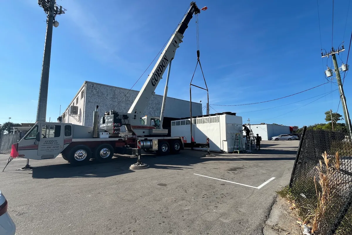 Gordon Crane's boom rigged over a long white packaged air-conditioning unit on a lowboy trailer, its row of condenser fans along the top and louvred coil panels down the side, with two crew on ladders working alongside it at a commercial building.
