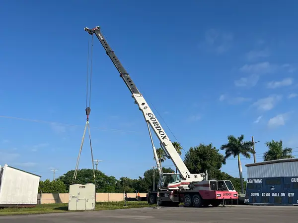A packaged rooftop air-conditioning unit hanging square in nylon slings from Gordon Crane's hook, the white boom raised above it and a paved lot behind.