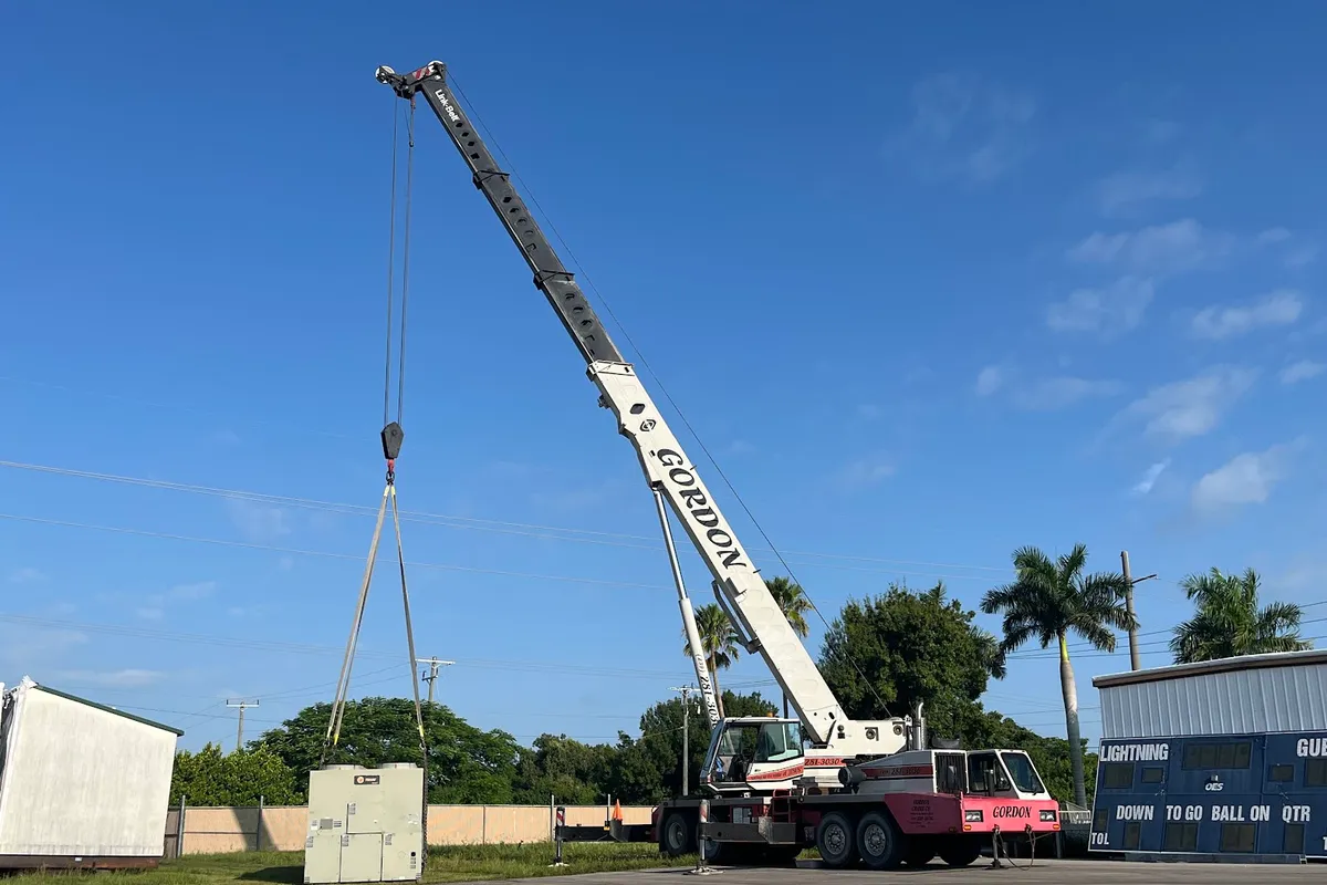 A packaged rooftop air-conditioning unit hanging square in nylon slings from Gordon Crane's hook, the white boom raised above it and a paved lot behind.