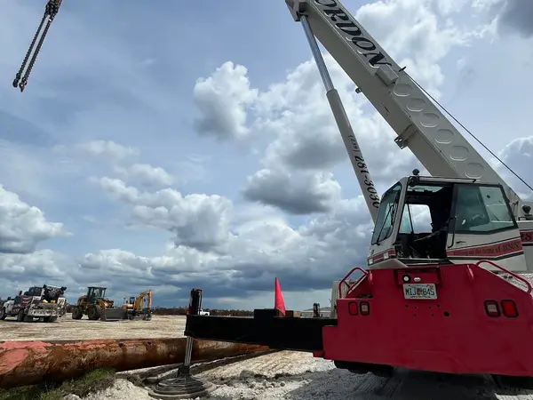 A red and white Gordon Crane rough-terrain machine on outriggers with its boom raised and rigging hanging, a length of large-diameter pipe on the fill beside it.