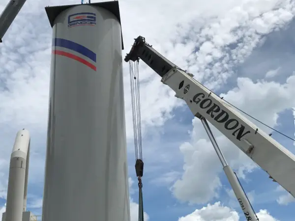 A white GENCOR silo hanging in slings between two crane booms, Gordon Crane's boom on the right, against a blue sky with broken cloud.