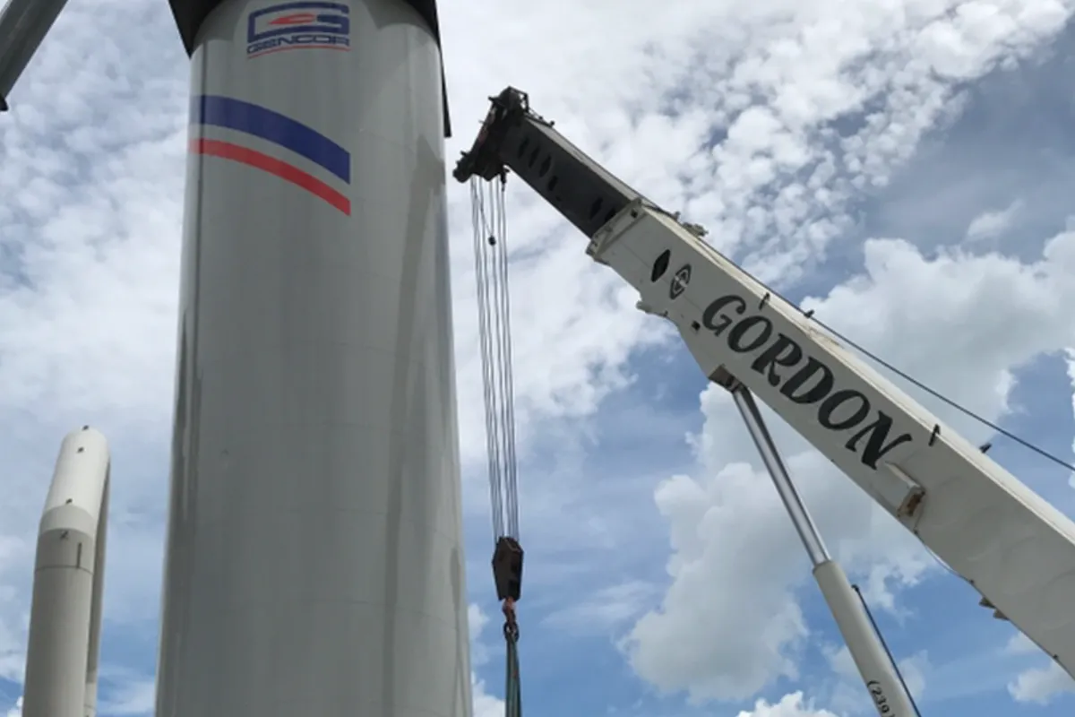 A white GENCOR silo hanging in slings between two crane booms, Gordon Crane's boom on the right, against a blue sky with broken cloud.