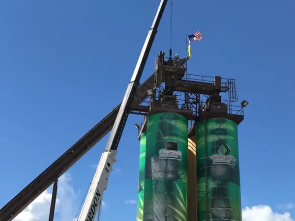 A Link-Belt crane in Gordon Crane livery with its boom extended to the top of two green-wrapped silos, flags flying from the platform.