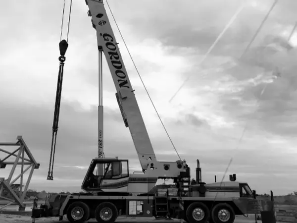 Black and white photograph of a Gordon Crane truck crane with its boom raised beside a steel frame section on open ground.