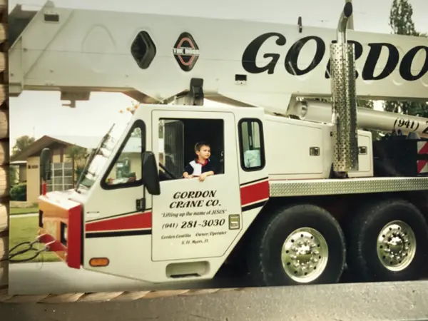 A young boy sitting in the cab of a Gordon Crane truck crane, the door lettered “Gordon Crane Co. — Lifting up the name of JESUS — (941) 281-3030 — Gordon Centilla, Owner/Operator, N. Ft. Myers, Fl”.